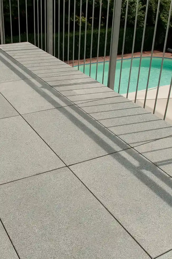 Close-up of light gray concrete tiles on an outdoor patio next to a metal fence with a swimming pool in the background.